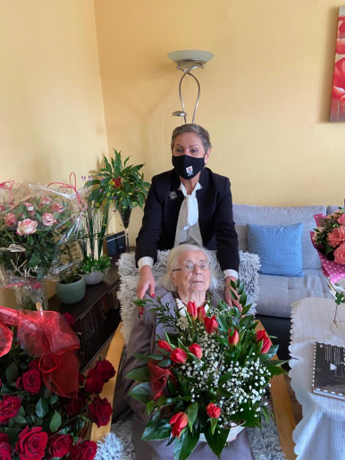 Na zdjęciu widzimy dwie kobiety - jedna siedzi na fotelu, druga stoi za nią i obejmuje ją za ramiona. Widać też dużo kwiatów, bukiety róż, tulipanów i innych pięknych kolorowych kompozycji.