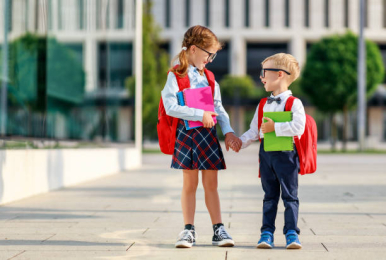 Na zdjęciu widzimy chłopczyka i dziewczynkę trzymających się za ręce, z kolorowymi zeszytami w drugich dłoniach i czerwonymi plecakami