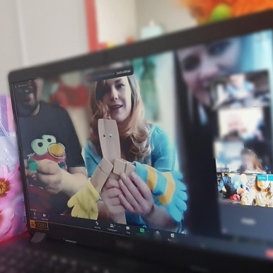 Na monitorze otwartego laptopa, młoda kobieta trzyma w dłoniach lalkę zrobioną z drewnianych łyżek kuchennych, ubraną w rękawiczki w kolorze niebiesko-żółtym , w tle mężczyzna  okienko z dziewczynka trzymająca drewniana łyżkę