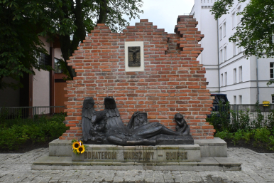 Pomnik Powstańców Warszawskich, obok bukier słoneczników, w tle szkoła, zieleń i inne budynki
