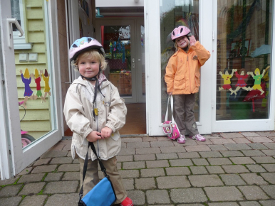 Na zdjęciu widzimy chłopca i dziewczynkę w kaskach i z torebkami stojący przed drzwiamy przedszkola.