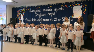 Na granatowym tłe widzimy napis uroczystość nadania imienia "Gryfickie Smyki" Przedszkolu Miejskiemu nr 21 w Słupsku oraz śpiewające dzieci i nauczycielki.
