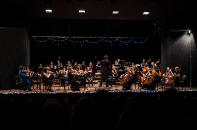 na scenie Filharmonii młodzi muzycy z instrumentami  podczas koncertu siedzą na krzesłach, tyłem  do widzów stoi dyrygent . Świąteczny nastój, światełka