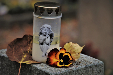 Znicz z obrazkiem aniołka postawiony na kamiennym obiekcie, obok liść i zaschnięty kwiatek