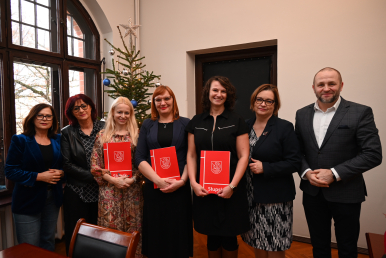 Wspólna fotografia nauczycieli odbierających akty mianowania, trzymający czerwone teczki Słupska w tle choinka