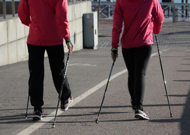 Obraz Sirpa P z Pixabay. Obraz przedstawiający dwie osoby spacerujące po asfalcie z kijami do nordic walking. Widoczne tylko nogi, tłów i ręce.