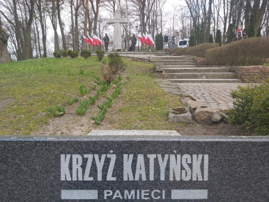 Na zdjęciu widzimy na pierwszym planie napis KRZYŻ KATYŃSKI PAMIĘCI. w tle schody prowadzące do pomnika Ofiar Katyńskich (krzyża) oraz dwóch żołnierzy i flagi Rzeczypospolitej Polskiej