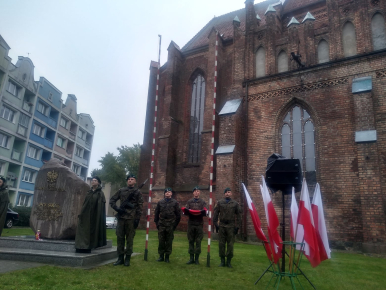 Na zdjęciu widzimy 4 żołnierzy i harcerkę; jeden z żołnierzy trzyma flagę, jeden broń; obok widzimy pomnik Polskiego Państwa Podziemnego, w tle kościół, a po prawej stronie flagi Polski