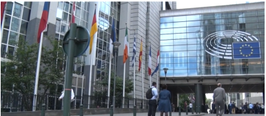 Na zdjęciu widzimy budynek, na którym znajduje się Flaga UE, obok na masztach flagi innych państw. Na chodniku dużó ludzi.