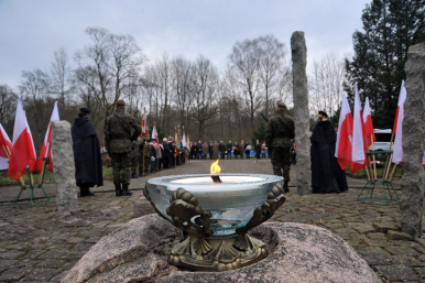 Na pierwszym planie płonący duży znicz stojący na kamieniu, na drugim planie uczestnicy oficjalnych uroczystości, wojsko, osoby duchowne, po obu stronach widoku flagi Rzeczypospolitej Polskiej