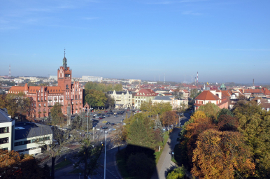 Na zdjęciu widać ratusz, pl.Zwcycięstwa z zaparkowanymi autami, kilka kamienic i budynki w tel oraz zieleń miejską