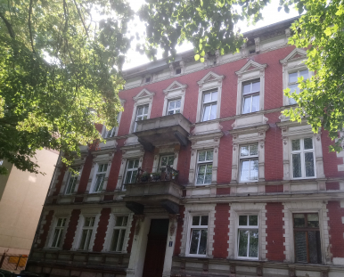Zdjęcie przedstawia budynek, w którym znajduje się lokal mieszkalny na sprzedaż. Dwupiętrowy budynek z czerwonej cegły, pojedyncze brązowe drzwi po środku budynku, nad nimi dwa balkony, obok drzwi tabliczka z numerem bloku 5