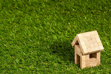 grafika prezentująca mały domek złożony z drewnianych klocków stojący na trawie