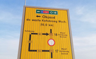 Przykładowa tablica wskazująca objazdy, w tle jezdnia, rondo, znaki drogowe, zieleń, drzewa