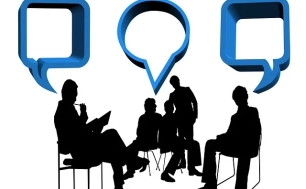 Na zdjęciu cztery osoby siedzą na krzesłach, jedna osoba stoi nad nimi "chmurki" dialogowe