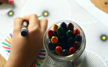 Na zdjęciu widoczna ręka dziecka, które koloruje kolorowankę. W pojemniku świecowe kredki