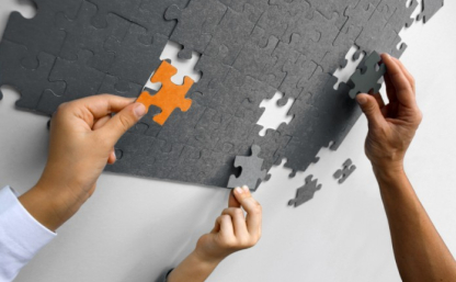 Grafika przedstawia trzy dłonie układające szare puzzle, jedna dłoń dokłada pomarańczowy element