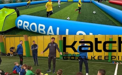 Boisko z dmuchanymi bandami i trenerzy rozmawiający z dziećmi.