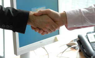 Na obrazku widoczne dwie dłonie mężczyz, którzy sobie je podają; w tle komputer i telefon