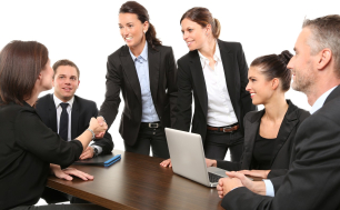 grupa mężczyzn i kobiet w garniturach przy stole z laptopem dwie kobiety, ściskaja sobie dłonie na powitanie