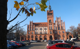 Na zdjęciu widzimy ratusz , przed nim plac z zaparkowanymi samochodami. Po lewej i prawej stronie drzewa.