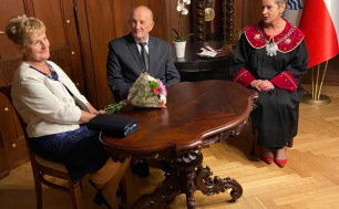 Na zdjęciu widzimy Prezydentkę Miasta oraz Jubilatów siedzących przy stole; na stole leżą kwiaty, w tle widać stojący zegar oraz flagi: Miasta Słupska oraz Rzeczypospolitej Polskiej