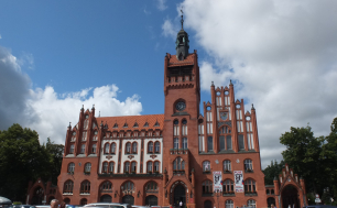 Na zdjęciu widzimy budynek ratusza z czerwonej cegły, po bokach zielone drzewa, przed ratuszem plac, kilku ludzi i kilkanaście zaparkowanych aut