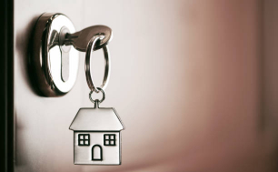 Na zdjęciu widzimy klucz do domu z brelokem domowym w dziurce od klucza w drzwiach