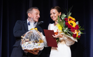 Pani Kasia Filipak otrzymuje nagrodę, w ręku trzyma teczkę z pismem, upominek, kwiaty, a mężczyzna obok niej wręcza kosz z upominkami