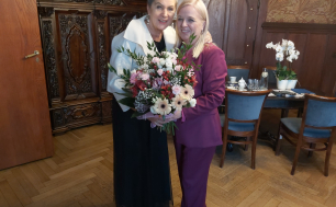 Prezydentka Słupska i Iwona Gozdek trzymające kwiaty, w tle stół z filiżankami, serwisem kawowym, szafa, meble gabinetowe