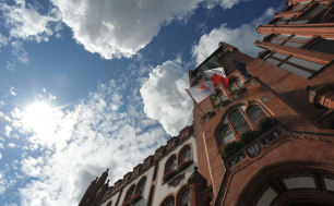 budynek ratusza po prawej stronie, po lewej widoczne niebieskie niebo z chmurami, na ratuszu zawieszona flaga Polski i flaga z herbem Słupska. Zdjęcie zrobione od dołu