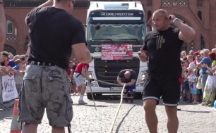 Na zdjęciu widzimy na pierwszym planie dwóch mężczyzn, trzeci trochę w tle ciągnie tira. Po bokach stoją widzowie obserwujący zmagania sportowca, a w tle widać słupski ratusz