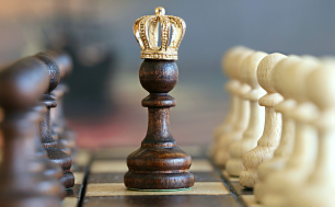 Zdjęcie szachownicy: w centrum czarny pion z ze złotą koroną, po prawej stronie rząd białych pionów, po lewej rząd czarnych pionów