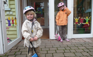 Na zdjęciu widzimy chłopca i dziewczynkę w kaskach i z torebkami stojący przed drzwiamy przedszkola.