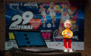 Na pierwszym planie rozłożony laptop, obok stojąca figurka Jurka Owsiaka wykonana z włóczki w tle plakat z napisem 29. Finał Wielkiej Orkiestry Świątecznej Pomocy