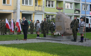 Pomnik Państwa Podziemnego w postaci kamienie/obelisku przed kościołem; obok żołnierze, prowadzący uroczystość , flagi RP, harcerze. W tle bloki, auta.