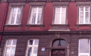 Zdjęcie przedstawia budynek, w którym znajduje się lokal mieszkalny na sprzedaż. Budynek z czerwonej cegły, brązowe podwójne drzwi wejściowe oraz tabliczka z numerem budynku 21 C.