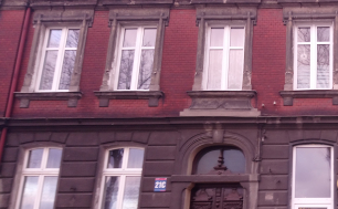 Zdjęcie przedstawia budynek, w którym znajduje się lokal mieszkalny na sprzedaż. Budynek z czerwonej cegły, brązowe podwójne drzwi wejściowe oraz tabliczka z numerem budynku 21 C.