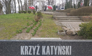 Na zdjęciu widzimy na pierwszym planie napis KRZYŻ KATYŃSKI PAMIĘCI. w tle schody prowadzące do pomnika Ofiar Katyńskich (krzyża) oraz dwóch żołnierzy i flagi Rzeczypospolitej Polskiej