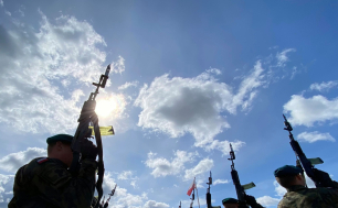 Na zdjęciu widzimy żołnierzy, którzy w górę kierują broń, w tle flaga Polski. Widać też niebo oraz chmury i słońce.