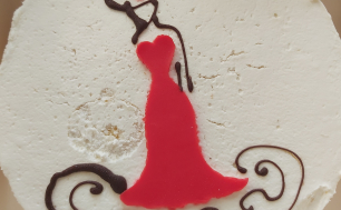 Na zdjęciu widzimy okrągły tort, biały, przyozdobiony w kształt kobiety ubranej w czerwoną sukienkę