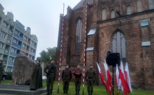 Na zdjęciu widzimy 4 żołnierzy i harcerkę; jeden z żołnierzy trzyma flagę, jeden broń; obok widzimy pomnik Polskiego Państwa Podziemnego, w tle kościół, a po prawej stronie flagi Polski