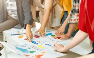 Widoczne ręce grupy osób skupionych przy stole roboczym, na którym leża są kolorowe kartki projektowe