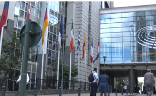 Na zdjęciu widzimy budynek, na którym znajduje się Flaga UE, obok na masztach flagi innych państw. Na chodniku dużó ludzi.