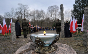 Na pierwszym planie płonący duży znicz stojący na kamieniu, na drugim planie uczestnicy oficjalnych uroczystości, wojsko, osoby duchowne, po obu stronach widoku flagi Rzeczypospolitej Polskiej