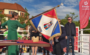 Na zdjęciu widzimy Panią Prezydent w zielonej sukience która trzyma sztandar skierowany w stronę klęczącego mężczyzny odbierającego w ten sposób symbolicznie z rak przedstawicielki władz samorządowych